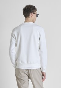 Sudadera blanca de manga larga, con puños acanalados y cuello redondo. Textura suave con un sutil detalle de logo en la parte trasera. Combinada con pantalones beiges.