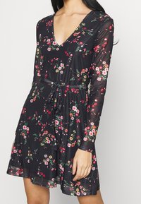 Schwarzes Blumenkleid mit langen Ärmeln, V-Ausschnitt und Bindegürtel. Es zeigt ein Muster aus Rosen und grünen Blättern auf einem durchsichtigen Stoff.