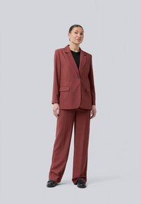 Blazer color ruggine e pantaloni a gamba larga coordinati, realizzati in tessuto texturizzato. Il blazer presenta un singolo bottone e due tasche frontali. Sotto, una maglietta nera.
