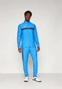 Lyseblå treningsdress i glatt stoff, med en marineblå horisontal stripe over brystet og svarte sideaksenter. Hvite joggesko.