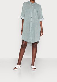 Abito camicia verde chiaro, con maniche corte, in tessuto texturizzato, lunghezza al ginocchio, dotato di taschino sul petto e con risvolti. Indossato con sandali bianchi.