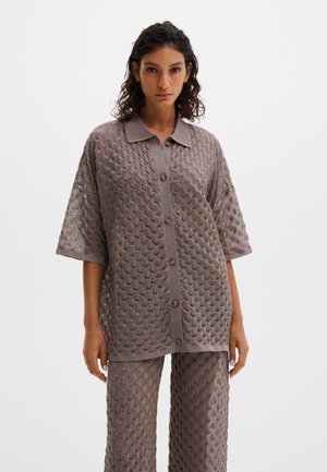 Femeie îmbrăcată într-un cardigan scurt cu nasturi, din tricot texturat taupe, cu mâneci scurte, și pantaloni largi asortați, stând în picioare pe un fundal alb simplu.