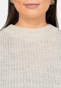 Lichtbeige gebreide trui met een losse, open weefstructuur en een geribbelde ronde hals. Accenten zijn onder meer textuurpatronen en zichtbare stiksels.