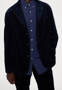 Mörkblå kordflossojacka med vågig krage, tre knappar och sidofickor, lagd över en mörkblå skjorta med prickar. Texturerad tygdetalj.