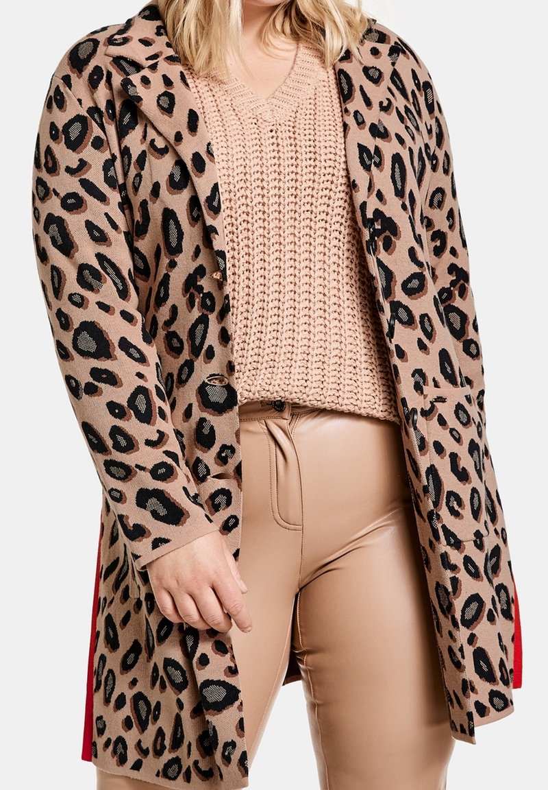 Cardigan con stampa leopardata su base beige, maniche lunghe e colletto. Indossato sopra un maglione rosa a maglia texturizzata e pantaloni beige aderenti.