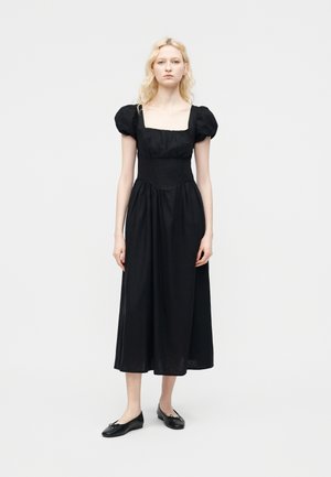 Donna in piedi che indossa un abito midi nero con maniche a sbuffo e scollo quadrato, abbinato a ballerine nere, su uno sfondo semplice.