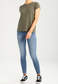 Oliwkowa koszulka z krótkim rękawem, luźny krój, zestawiona z dopasowanymi jasnoniebieskimi dżinsami i czarnymi trampkami z białą podeszwą. Proste tło.