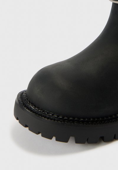 Černá kotníková bota z gumy s kulatou špičkou, texturovaným povrchem a silnou podrážkou s vzorkem a tkaným zdobením kolem paty.
