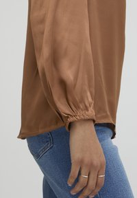 Blusa di satin marrone con maniche lunghe e a sbuffo; presenta polsini arricciati e una texture liscia e lucida; abbinata a jeans in denim blu.