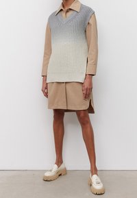 Gilet en tricot à col en V avec un dégradé de nuances du gris au crème, porté sur une robe-chemise beige. Des mocassins blancs épais complètent la tenue.