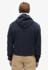 Man met de rug naar de camera, gekleed in een navyblauwe hoodie over een wit shirt en beige broek, tegen een effen witte achtergrond.