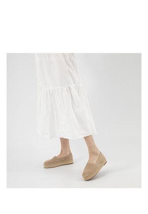 Person trägt beige Espadrille-Ballerinas kombiniert mit einem weißen, luftig-fließenden Kleid bis zum Knöchel vor einfachem Hintergrund.