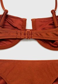Tmavo hrdzavý bikiny top so nastaviteľnými popruhmi, jemnou látkou a uzáverom na zadnej strane. Má hladké, bezšvové okraje.