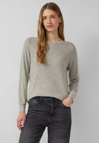 Kobieta z długimi blond włosami, ubrana w szary wzorzysty top z długim rękawem i czarne dżinsy, stoi z jedną ręką w kieszeni na jednolitym tle.
