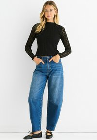 Black knitted long-sleeve top with a textured pattern paired with high-waisted blue denim jeans. Black ballet flats complete the look.