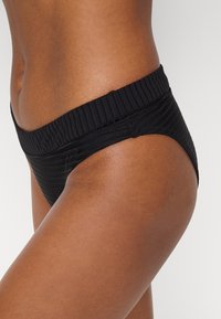 Fundo de bikini preto com nervuras, apresentando uma faixa larga texturizada na cintura, com um corte baixo e bordas lisas para conforto e ajuste.