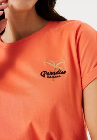 Korala bawełniana koszulka z krótkim rękawem, zdobiona haftowanym złotym i czarnym napisem "Paradise Garcia" oraz wzorem palmy.