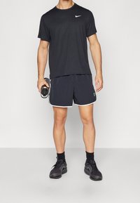 Černé tričko Nike s krátkými rukávy, spárované s tmavými šortkami s bílými akcenty a černými sportovními botami. Lahvička na vodu držena v ruce.