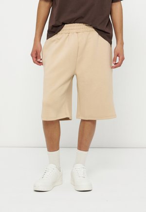 Άνδρας που φοράει μπεζ σορτς μέχρι το γόνατο, άσπρες κάλτσες, άσπρα αθλητικά παπούτσια και καφέ μπλουζάκι, στέκεται μπροστά σε λευκό φόντο.