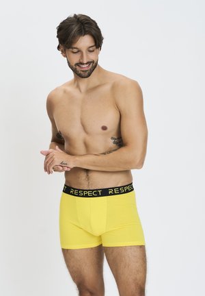Žlté bavlnené boxerky s čiernym pásom s nápisom "RESPECT." Tkanina vyzerá hladko a tesne sa prispôsobuje pásu a stehnám.