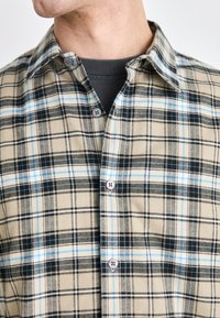 Tan plaid flannel shirt featuring black and light blue lines, with button-down collar and two-button placket in contrasting colors.