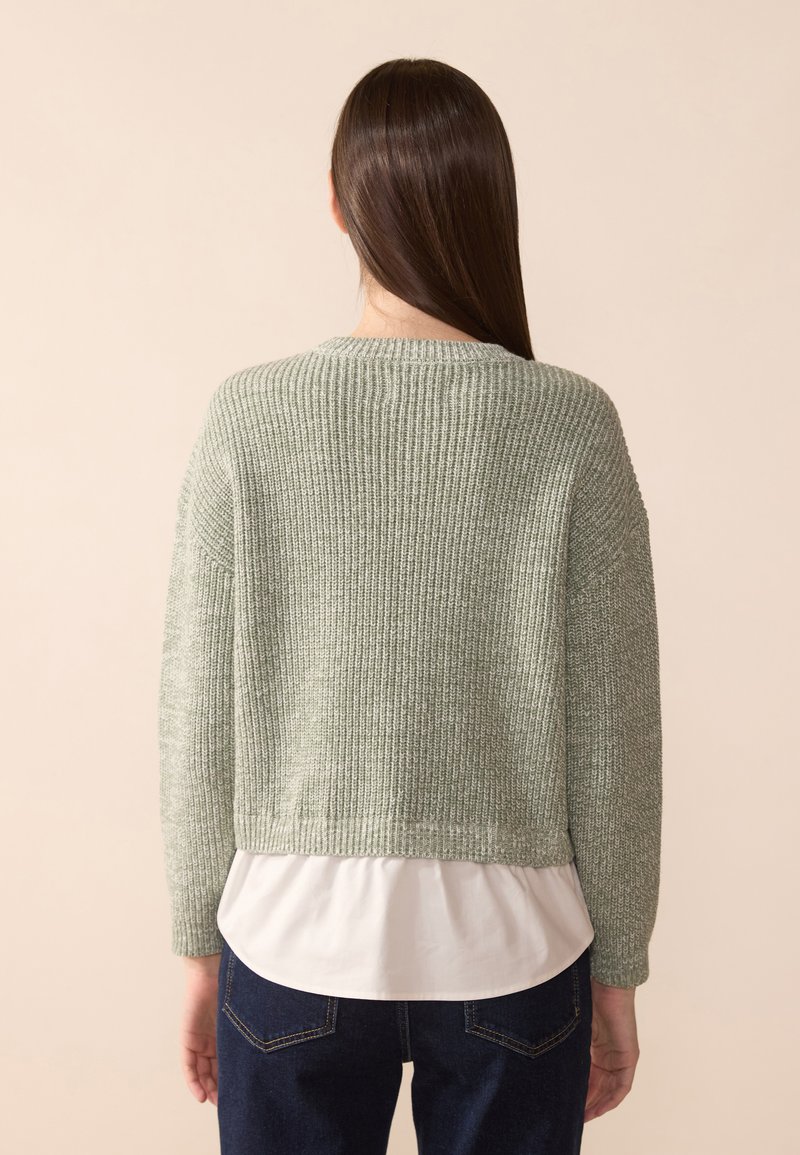 Pull en tricot vert clair avec une coupe ample, texture côtelée et manches longues, superposé sur une chemise blanche, accompagné de jeans foncés.
