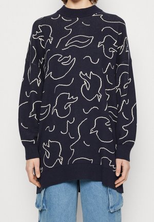 Marineblauer übergroßer Pullover mit weißen abstrakten Vogelmuster, mit geripptem Kragen, Bündchen und Saum, getragen über hellblauen Cargo-Jeans.