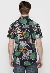 Camisa de manga corta y botones en color navy, con un patrón de hojas tropicales en verde, amarillo y beige, hecha de una tela ligera.