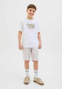Vit kortärmad t-shirt med "JACK & JONES"-logotyp, ljusbeiga shorts, vita strumpor och beiga skor, stående mot en enfärgad bakgrund.
