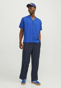 Chemise à boutons à manches courtes bleue associée à un pantalon cargo marine et à des baskets bleues. Le tissu semble léger avec une texture lisse.