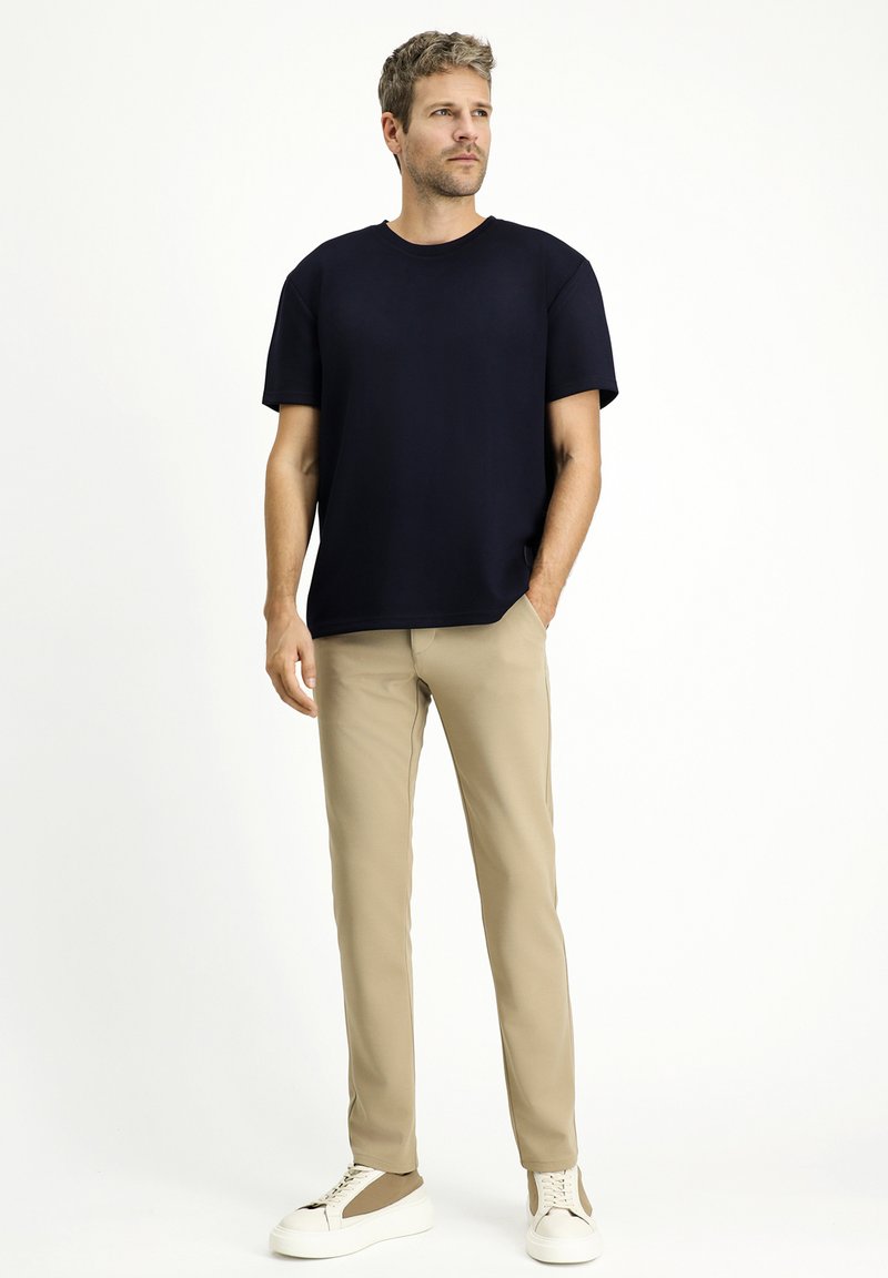 T-shirt nera a maniche corte abbinata a pantaloni beige slim-fit e sneakers bianche con dettagli marroni. Taglio ampio, tessuto morbido e design semplice.