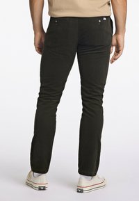 Donkergroene corduroybroek met een slim fit, voorzien van achterzakken met knoopsluitingen en een subtiele textuur. Draagt met witte canvas sneakers.