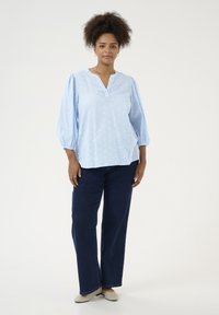 Blouse bleu clair avec un décolleté en V, texturée avec des motifs en relief et des manches volumineuses. Associée à un pantalon en jean large bleu marine et des chaussures beiges.