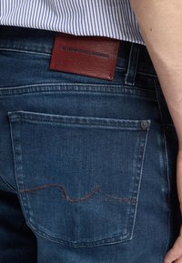 Jeans in denim blu scuro con una finitura testurizzata, dotati di un'etichetta in pelle marrone del marchio e dettagli di cucitura sottili sulla tasca posteriore.