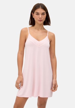 Mujer que lleva un camisón sin mangas de color rosa claro con tirantes finos y ribete de encaje en el escote, de pie frente a un fondo liso.