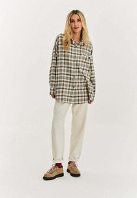 Blondynka stoi, ubrana w oversize'ową szarą i brązową koszulę w kratę, kremowe spodnie podwinięte przy nogawkach, czerwone skarpetki i beżowe buty turystyczne na białym tle.