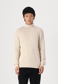 Sweater bege canelado com gola alta e mangas longas; apresenta um corte descontraído, combinado com calças escuras.