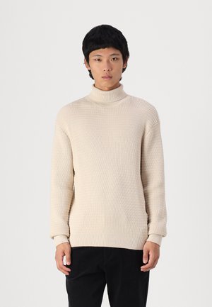 Beige, ribbad stickad polo med långa ärmar; har en hög krage och avslappnad passform, i kombination med mörka byxor.