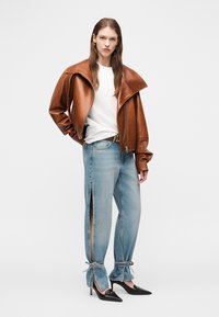 Frau in brauner Lederjacke, weißem T-Shirt, hellblauen Jeans mit seitlichen Schlitzen, die an den Knöcheln gebunden sind, und schwarzen spitzen High Heels, stehend vor weißem Hintergrund.