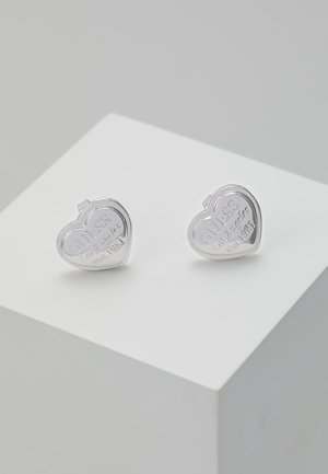 Boucles d'oreilles clous en argent en forme de cœur gravées "GUESS Los Angeles Est. 1981" sur un socle blanc sur fond gris.