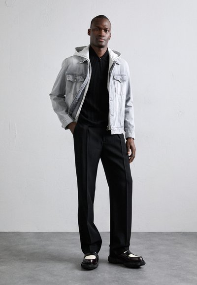 Grijze denimjack met een capuchon, zwarte geribde trui, zwarte broek en chunky schoenen met een glanzende accent. Eenvoudige achtergrond.