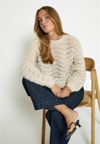 Cream knit sweater with a wave pattern, featuring a round neckline and ribbed cuffs, paired with dark denim flared pants and black sandals.