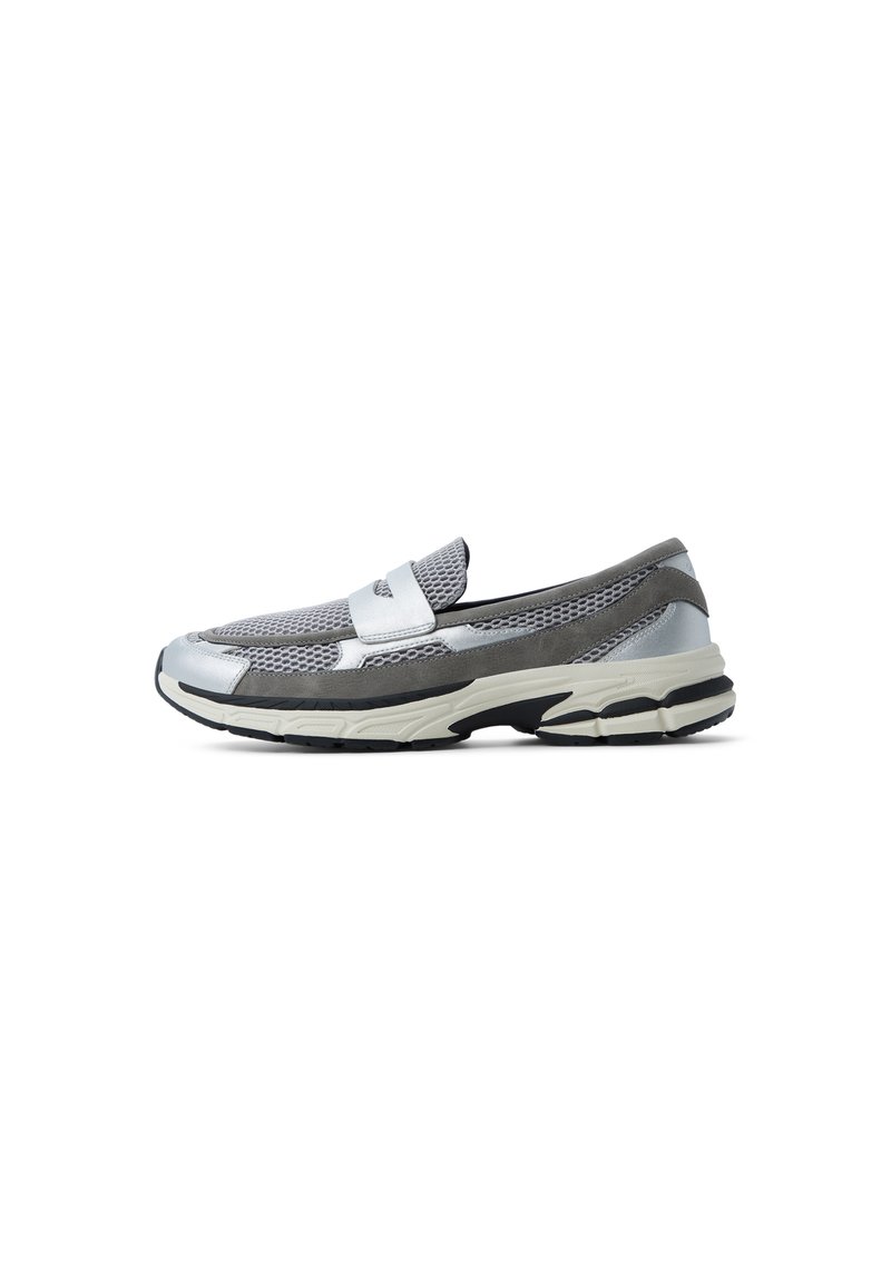 Zapatilla deportiva sin cordones con parte superior de malla gris, acentos plateados y una suela gruesa en blanco y negro. Cuenta con un collar acolchado y detalles de correa.