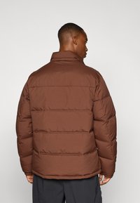 Braune, gepolsterte Jacke mit vertikalen Nähten, hohem Kragen und elastischen Bündchen. Glatte Stofftextur mit gestepptem Design.