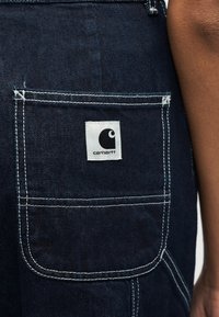 Τσέπη από σκουρόχρωμο τζιν με λευκή ραφή και λευκή ετικέτα με το λογότυπο Carhartt, φορεμένη από ένα πρόσωπο με μέτριο καφέ δέρμα ορατό στα δεξιά.