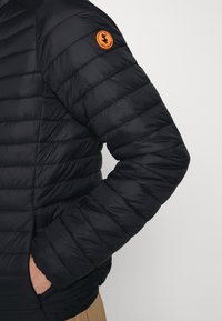 Schwarze Daunenjacke mit horizontalem Steppmuster. Verfügt über ein orangefarbenes, rundes Logo am linken Ärmel und einen Reißverschluss an den Seiten.