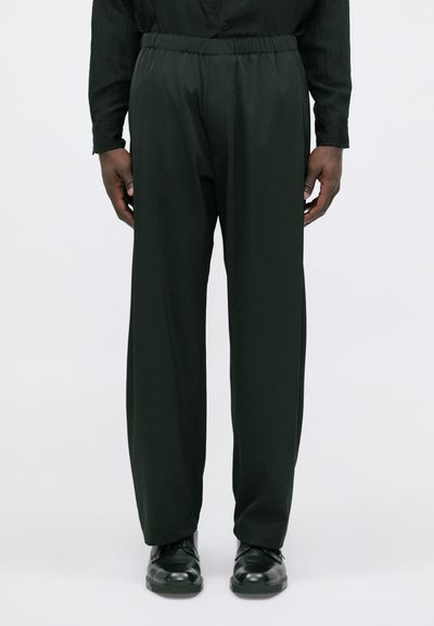 Bărbat îmbrăcat în pantaloni negri de costum cu talie elastică și pantofi negri de costum, stând pe un fundal simplu deschis.
