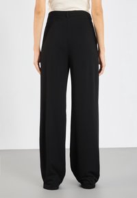 Femme portant un pantalon large taille haute noir se tenant devant un fond clair.
