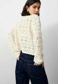 Pull à mailles creuses crème avec un ourlet en forme de vague et des manches longues, associé à un jean taille haute bleu foncé.
