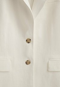 Gros plan sur un blazer de couleur crème avec deux boutons en écaille de tortue et des poches à rabat de chaque côté.